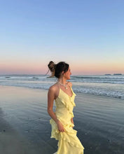 Load image into Gallery viewer, A woman standing on a beach at sunset wearing a yellow maxi dress with ruffled detailing, with the ocean and a boat in the background.
