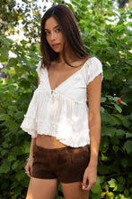 Load image into Gallery viewer, Woman wearing a white blouse and brown shorts standing in a green outdoor setting
