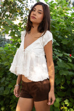 Load image into Gallery viewer, Woman wearing a white blouse and brown shorts standing in front of green foliage
