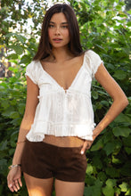 Load image into Gallery viewer, Woman wearing a white lace top and brown shorts standing in front of green foliage.
