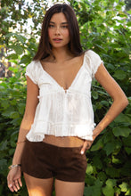 Load image into Gallery viewer, Woman wearing a white lace top and brown shorts standing in front of green foliage.
