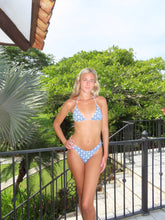 Load image into Gallery viewer, Woman in a blue patterned bikini standing outdoors with greenery and a metal railing in the background.
