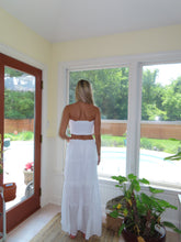Load image into Gallery viewer, Woman in a white dress standing in a room with large windows and a plant.
